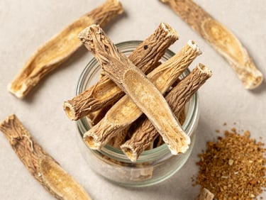 Overhead flat-lay of dried licorice root sticks and powdered extract in a small glass jar arranged on a pale linen cloth under bright diffused studio light, close-up framing showing root fiber texture