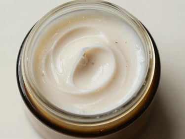 Extreme close-up overhead studio shot of a small open jar of herbal face cream on a neutral off-white surface, cream texture visible with fine botanical particulates, bright diffused daylight from above, no props