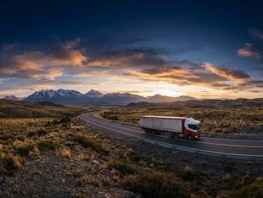Wide panoramic shot of a modern heavy-duty freight truck driving on a scenic highway in the Southern Cone at crisp morning daylight, with a low horizon and expansive sky, deep navy and industrial orange tones, wide-angle heroic lens.