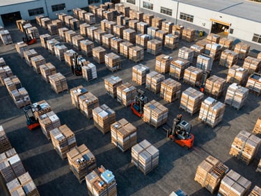Aerial view of a large, organized logistics warehouse with forklifts moving pallets, clean and efficient operation, crisp daylight, deep navy and industrial orange tones.