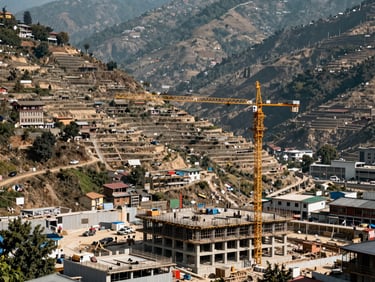 Wide-angle shot of a Kathmandu valley construction site at midday, terraced hillsides visible in background, crane and partially built concrete structure in foreground, natural light, no people posed — scale and progress evident