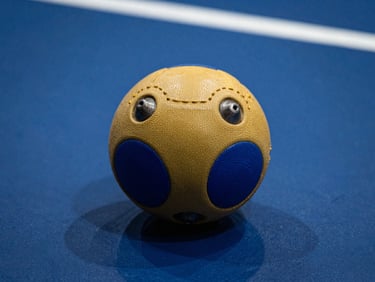 Close-up of a goalball with embedded bells resting on a high-contrast blue court, sharp directional light highlighting the tactile textures, 35mm lens.