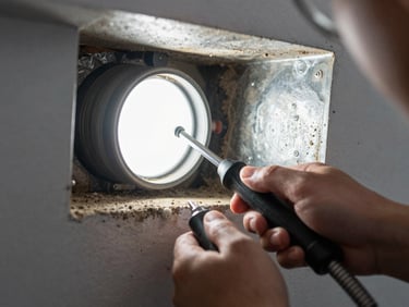 Close-up of technician hands using a flexible camera probe inside a residential air duct, bright LED illuminating dust-caked metal walls, tool grip visible in foreground