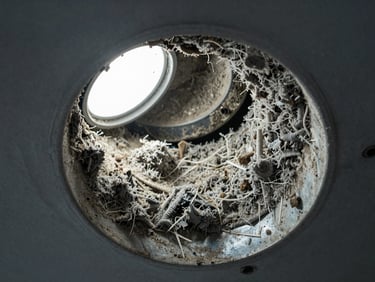 Close-up of a residential air duct interior showing accumulated dust, debris, and grey lint coating the metal walls, bright work light illuminating the blockage, shot from duct opening looking inward