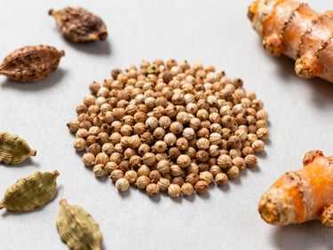 Bright editorial overhead flat-lay of sorted coriander seeds, cardamom pods, and turmeric roots on a clean light-grey surface, natural daylight, sharp focus