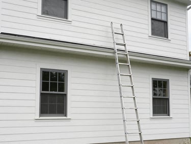 Wide shot of a two-story home exterior freshly painted in natural daylight, clean siding lines, new trim color visible, ladder resting against wall, no people