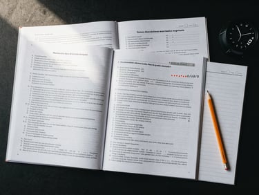 Wide flat-lay of a German language exam preparation kit spread across a dark desk surface: open workbook with practice test pages, answer sheet with pencil marks, a timer placed to the right edge, soft directional studio light from upper left, desaturated cool palette, no faces or legible text