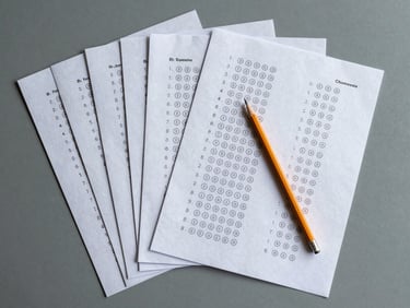 Flat-lay overhead of B1 exam practice papers fanned out on a grey surface, a pencil placed diagonally across them, one sheet showing multiple-choice answer bubbles, cool natural light, no text legible, muted tones