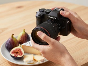 Close-up of hands holding a professional camera over a beautifully styled plate of figs and cheese, soft north-facing daylight, natural wood background