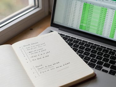 Overhead view of a notebook with handwritten financial model notes next to a laptop showing Excel rows with formula cells highlighted in green, daylight from a window