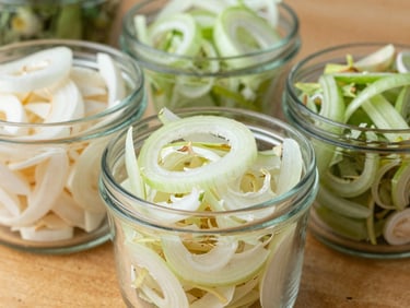 Close-up of dehydrated sliced onions and garlic flakes in clean industrial glass containers on a wooden table, bright daylight, sharp focus.