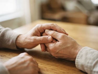 Close-up of supportive hands gently holding each other on a warm oak table, soft diffused window light, shallow depth of field, 35mm