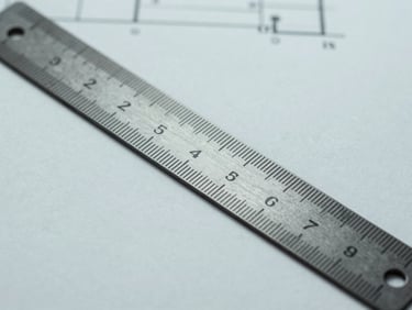 Macro photography of an architectural ruler and blueprint on a clean, desaturated background, high-contrast studio light.