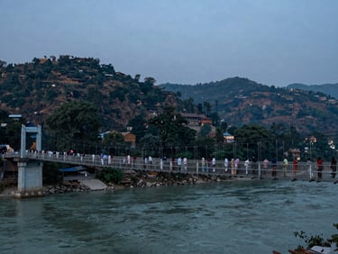 Wide documentary frame of Rishikesh suspension bridge at dusk, pilgrims crossing single-file, Ganga visible below through bridge gaps, blue-grey sky above forested hills, no artificial light