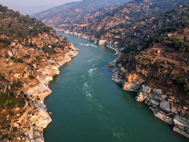 Aerial wide frame of Devprayag confluence, Alaknanda and Bhagirathi rivers meeting in a sharp V, deep blue-green water against ochre rock walls, forested ridges beyond, early morning light