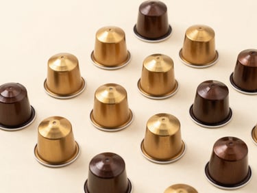 Overhead minimalist studio shot of neatly arranged coffee capsules in warm metallic gold and deep brown tones on a clean cream surface