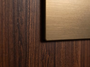 Extreme close-up of dark walnut paneling meeting a brushed bronze sensor plate, warm side lighting highlighting the rich wood grain and precise metal edge.