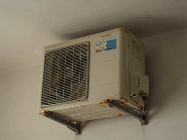 Old window AC unit wedged into a wall opening in a Noida flat, natural indoor light, visible rust on the casing edges, unit label partially faded