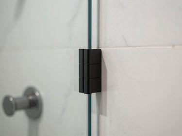 Close-up of a frameless glass shower enclosure joint, matte black hardware profile meeting a clean marble wall, natural diffused daylight, sharp focus on the mitred edge