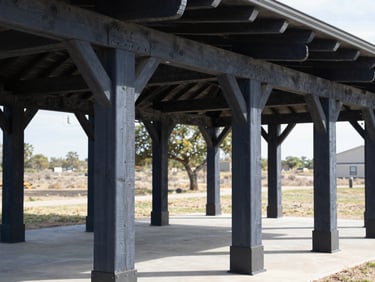 Heavy timber framing of an expansive outdoor pavilion, thick cedar posts bolted with black powder-coated steel brackets.