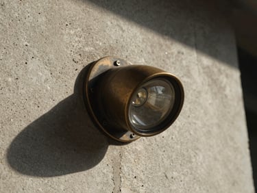 Macro photograph of a raw sand-cast bronze downlight fixture, harsh direct sunlight casting deep dramatic shadows on a textured concrete wall, warm earthy tones, 35mm film.