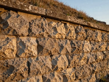 Detail of mortared retaining wall stone courses, golden hour side-lighting revealing material texture and tight joint work, slope visible behind the wall, no people