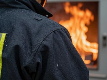 Structural firefighting gear undergoing flame engulfment testing in a clean laboratory thermal chamber, intense orange fire backlighting, sharp focus on fabric weave, 35mm.