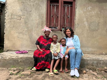A smiling group of two women and two children sitting outside a house in an African village.