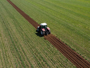 Wide aerial drone shot of a tractor cultivating a vast field, creating precise furrows, commercial agriculture photography, bright direct sunlight