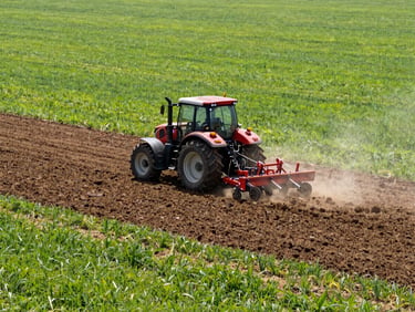 Heavy-duty tractor tilling rich soil in a vast field, dust rising under bright midday sun. Wide-angle lens, 35mm, commercial agriculture photography.