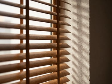 Close-up of light filtering through half-open wood Venetian blind slats, casting sharp horizontal shadow lines on a textured plaster wall.
