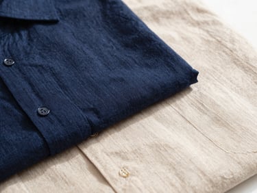 Bright editorial close-up of folded linen shirts in navy and beige, soft natural daylight, textured fabric weave visible, minimalist styling.