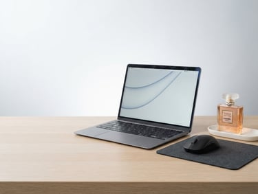 Bright high-key product photography of a neatly organized desk with a laptop, a perfume bottle, and a gaming mouse, clean studio light, crisp details, white and soft gray background, 50mm lens, expansive negative space to the left.