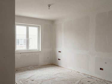 Residential room mid-renovation showing exposed plasterwork and fresh wall prep, daylight from a window, floor protected with dust sheets, shot at standing eye level from the doorway