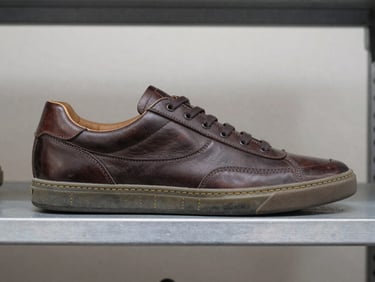 Side profile of a classic used leather sneaker on a clean metal shelf, bright neutral daylight, clear sole stitching detail.