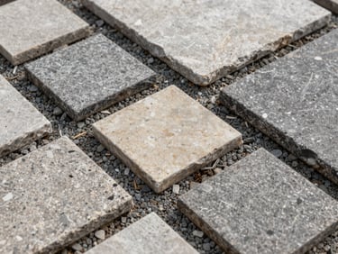 Overhead close-up of hardscape stone samples and pavers laid on dry aggregate, varied textures and natural tones, flat overcast daylight, no people, material detail framing
