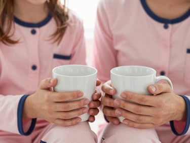 Close-up of mother and daughter hands holding warm mugs, wearing matching soft pink and navy blue pajamas, cozy morning light