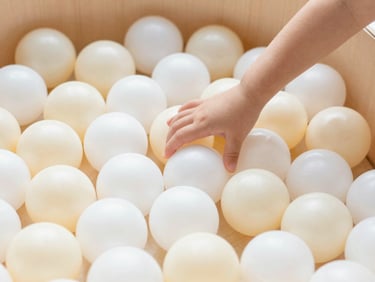 Close-up of a premium wooden ball pit filled with soft cream and pearl white balls, a child's hand gently reaching in, bright high-key lighting.