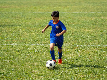 Candid shot of a young boy in a blue jersey dribbling a soccer ball on a sunny grass field, vibrant action