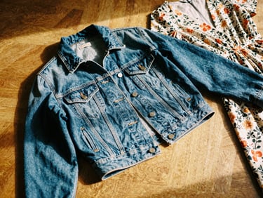 Flat lay of stylish women's clothing, including a vintage denim jacket and a floral dress, on a wooden floor in warm natural light, 35mm film aesthetic