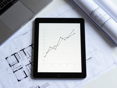 Overhead clean desk layout with architectural blueprints and a tablet displaying line charts, cool bright daylight, graphite tones.