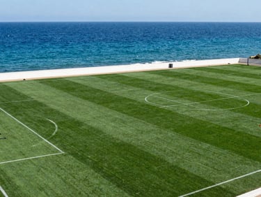 A scenic Greek coastal soccer pitch with deep matte forest green grass, overlooking the blue Mediterranean sea under bright natural sunlight, 35mm lens.