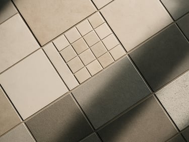 Overhead shot of ceramic and porcelain tile samples arranged on a showroom floor, varying grout widths visible, natural daylight casting soft shadows across the surface textures