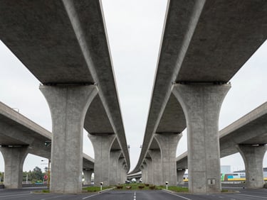 Symmetrical composition of concrete support pillars beneath a modern highway, clean lines, diffused overcast daylight.