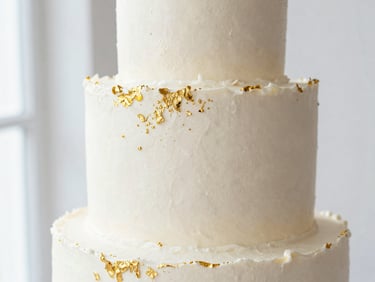 Macro close-up of a three-tiered wedding cake with textured white buttercream and delicate edible gold leaf, bright side-lighting, clean minimalist studio background.