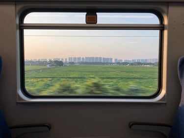 Wide shot through a bullet train window at dawn, blurred green farmland and distant city skyline passing by, warm early light on the glass, empty seat armrest in foreground
