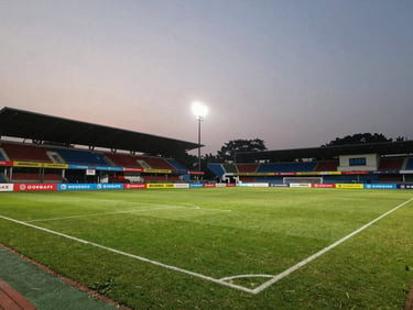 Wide environmental shot of a premium floodlit football turf in Kolkata, featuring high-visibility brand banners along the touchline, dramatic evening lighting.