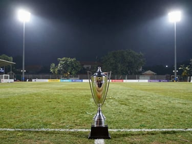 A sleek silver trophy standing on the touchline under dramatic evening floodlights, wide environmental shot of a premium Kolkata turf pitch.