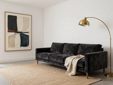 Wide environmental shot of a complete living room, right-bleed composition, deep charcoal velvet sofa with a natural flax-linen throw, a large-format abstract artwork on the wall, a brushed brass arc floor lamp, flat-weave rug in oat tones, bright even studio-quality daylight, full spatial arrangement visible, material detail clear, no clutter