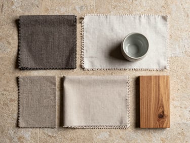 Overhead flat-lay composition of grouped furniture and material samples — fabric swatches, a small ceramic, a wood block — on a warm stone surface, even bright studio light, honest material rendering